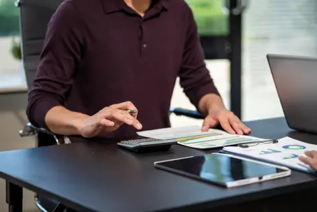 Businessman using a calculator and reviewing financial charts at an office desk with a tablet and laptop, planning a budget or checking expenses.