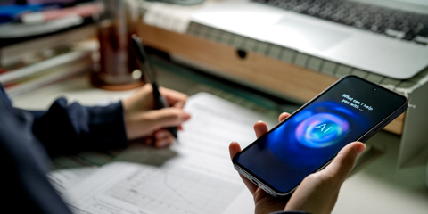 An Asian teenage student interacts with an AI chatbot on a smartphone while studying at a desk with a laptop, notes and stationery. The scene highlights modern learning and technology integration.