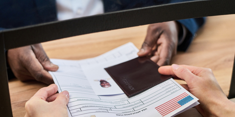 Unknown woman handing visa application form and passport to man behind counter at visa center, both sets of hands visible exchanging documents during official process