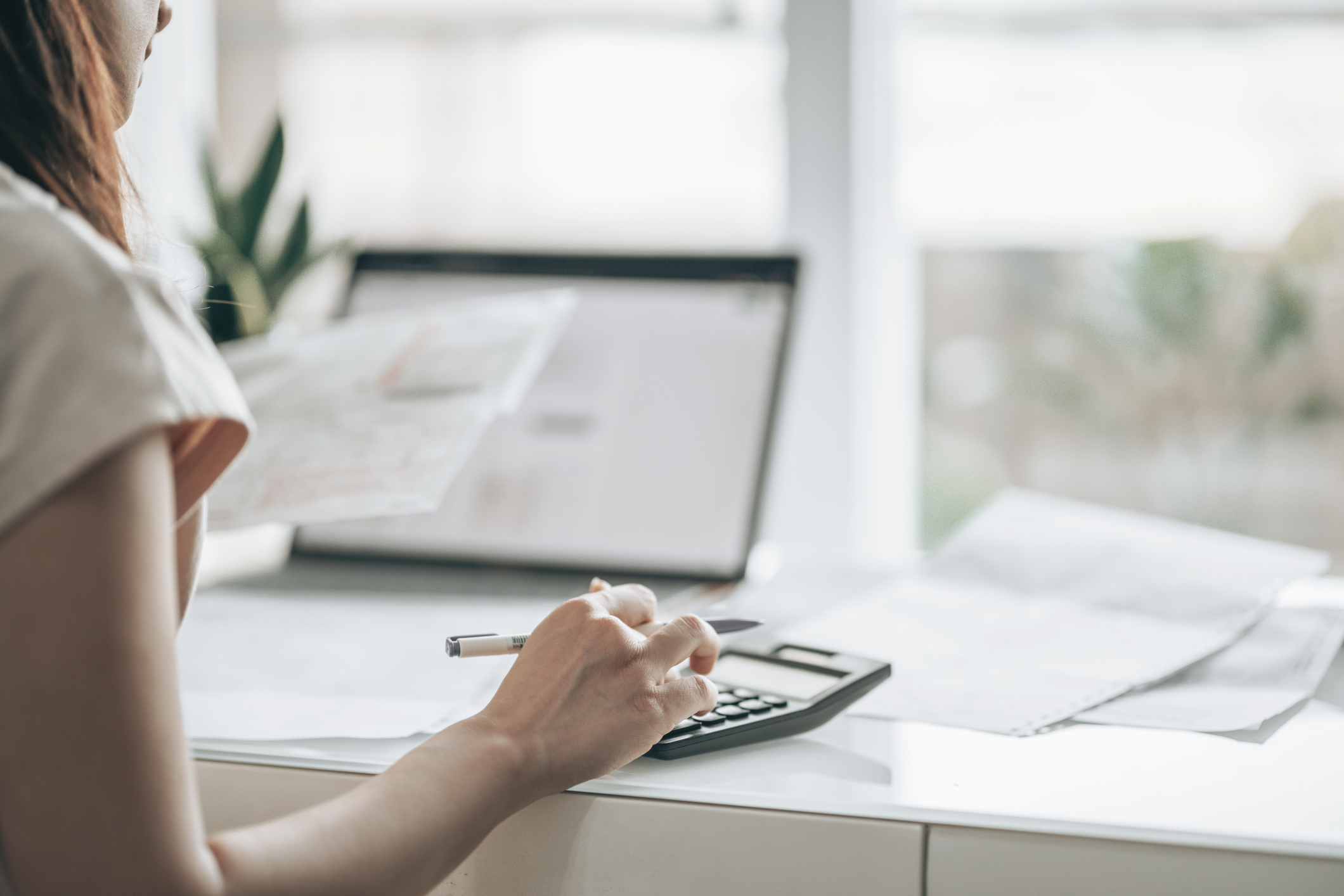 The person holds a pen poised over a calculator, surrounded by paperwork and a laptop displaying a tax form. The scene represents a busy, freelance workspace focused on financial tasks.