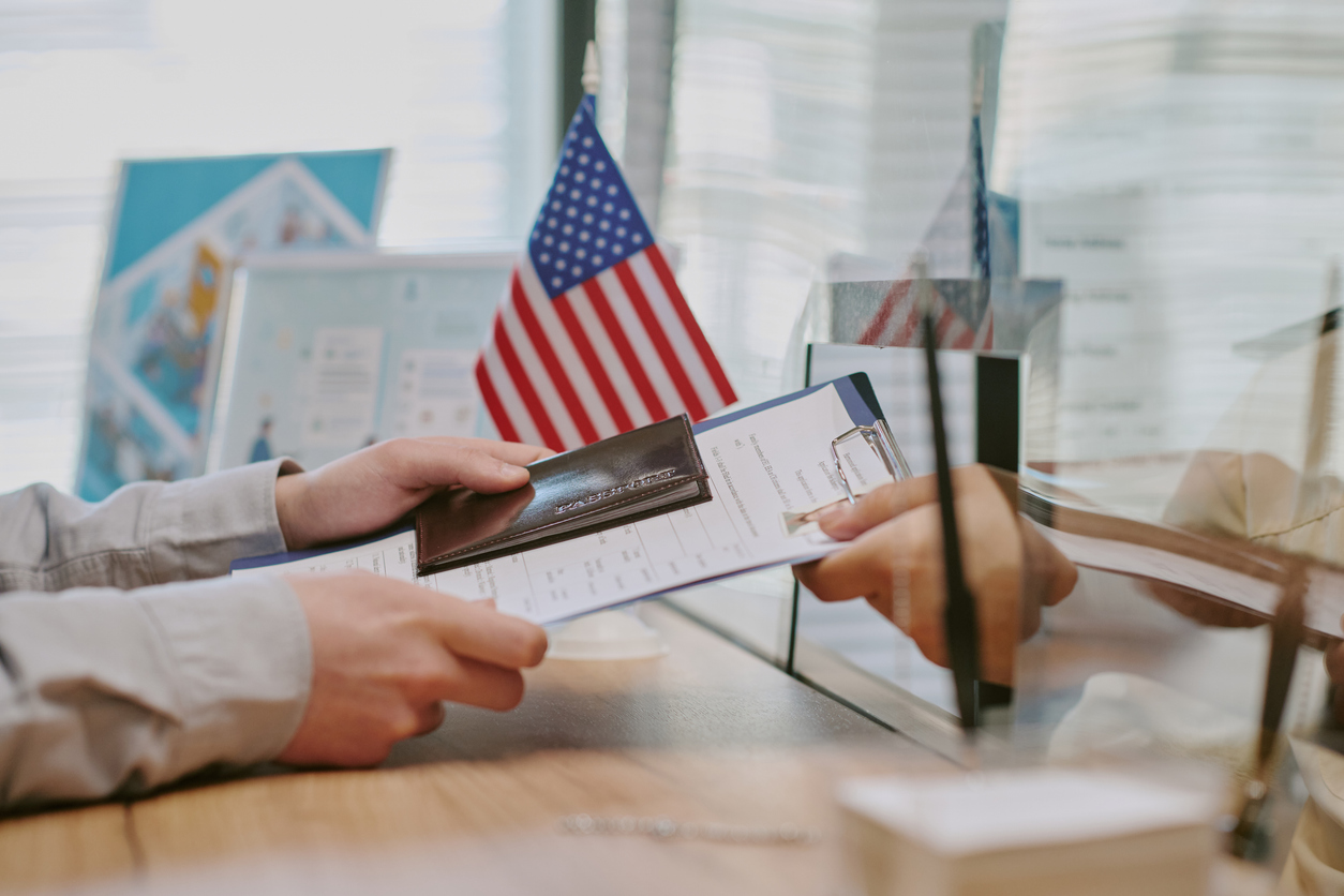 Hands holding passport and form while completing documentation on desk at visa center. Visible American flag and documents in background