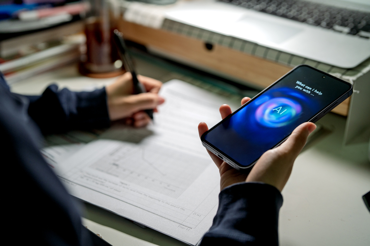 An Asian teenage student interacts with an AI chatbot on a smartphone while studying at a desk with a laptop, notes and stationery. The scene highlights modern learning and technology integration.