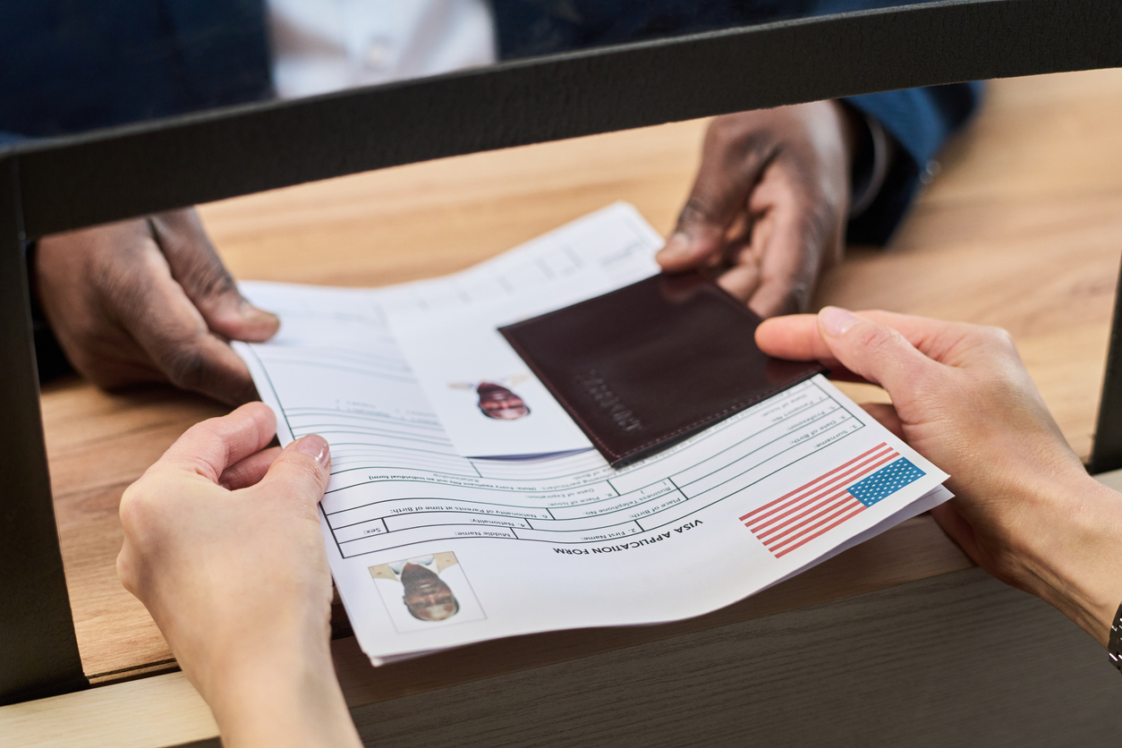 Unknown woman handing visa application form and passport to man behind counter at visa center, both sets of hands visible exchanging documents during official process